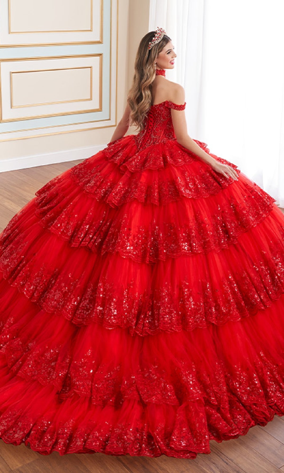 A woman in the Quinceanera Dress PR30181 By Princesa, featuring sparkling tiers and off-the-shoulder sleeves with a tiara, stands in a decorated room, facing away from the camera.